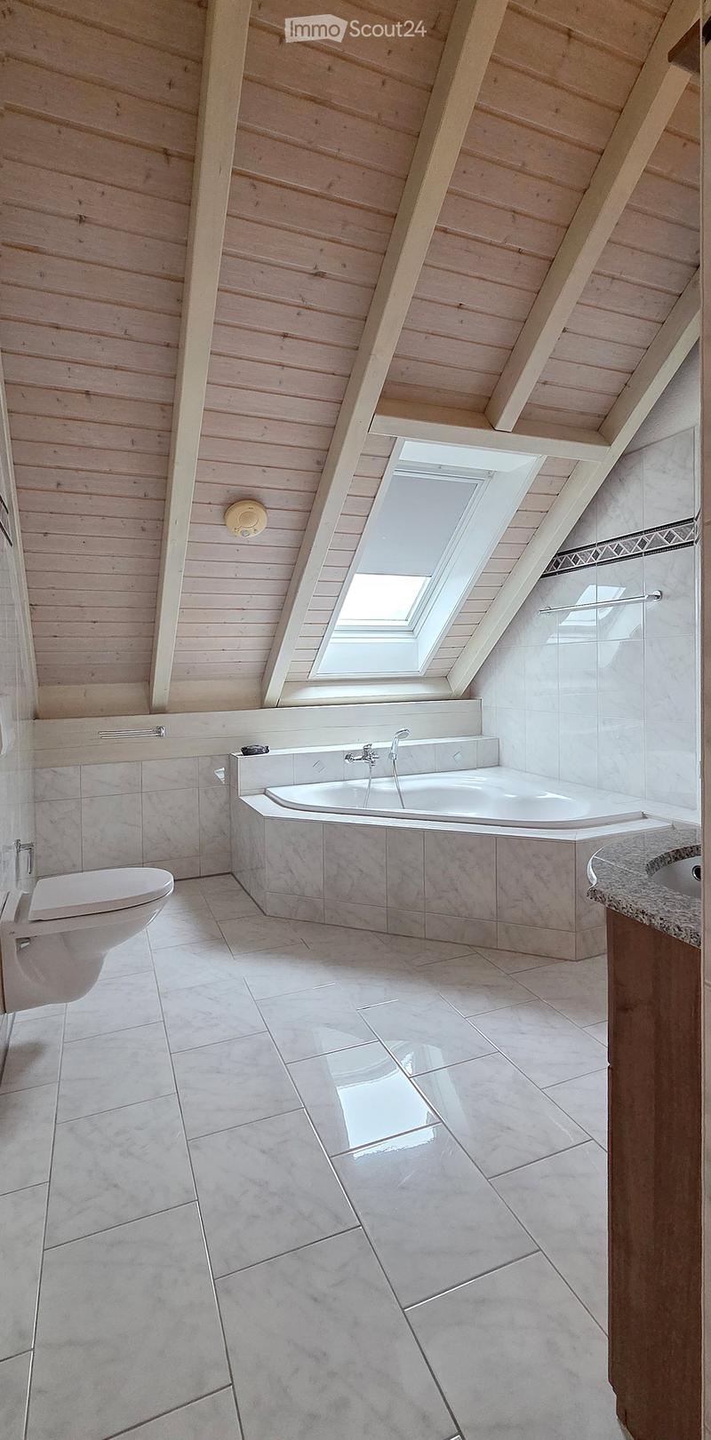 Modern bathroom with a large bathtub, toilet, sink, and a skylight window above the tub. The room features light wooden paneling on the ceiling and walls, and a tiled floor.