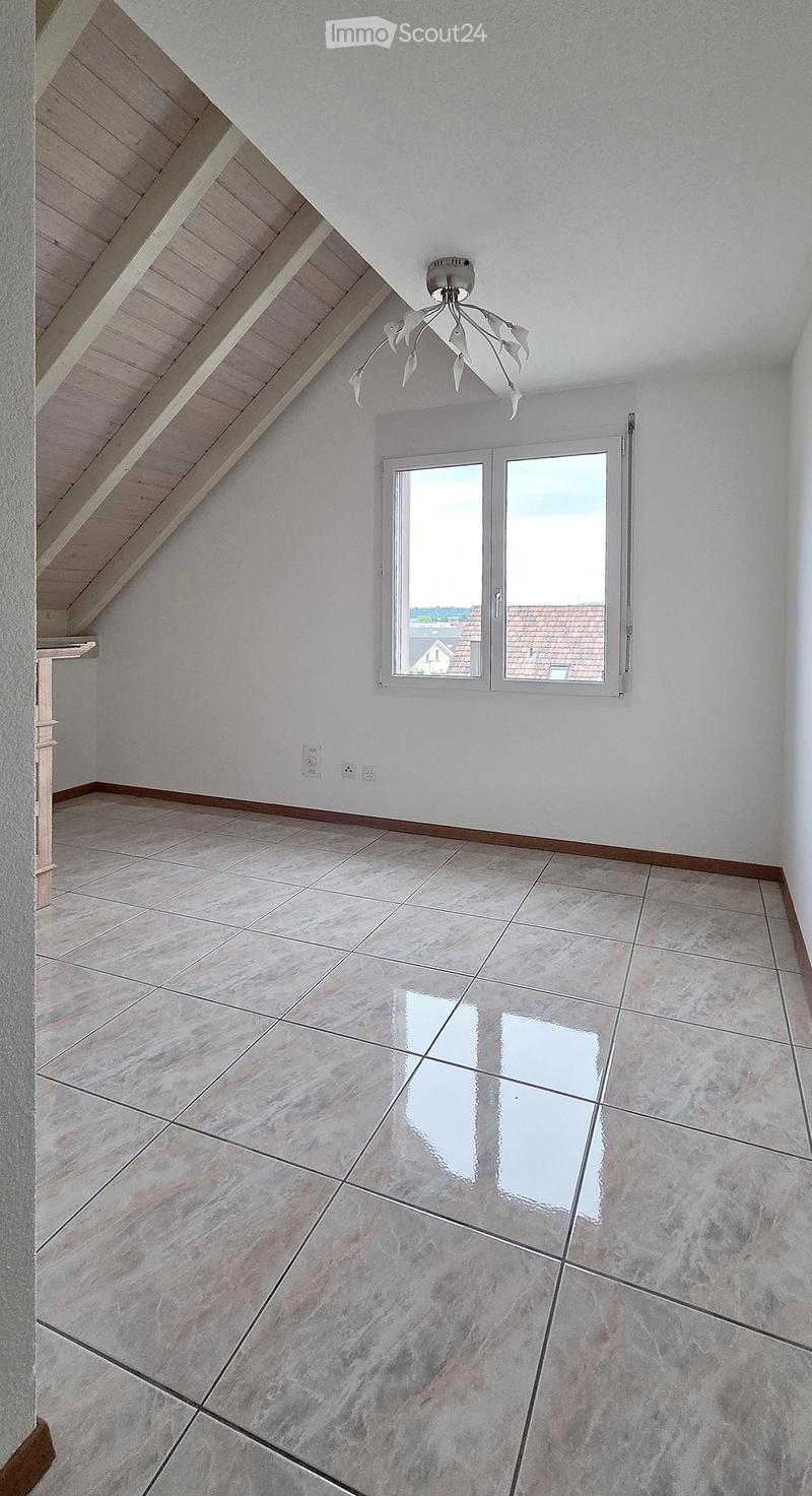 Empty attic room with wooden slanted ceiling, large window, tiled floor, and modern light fixture.