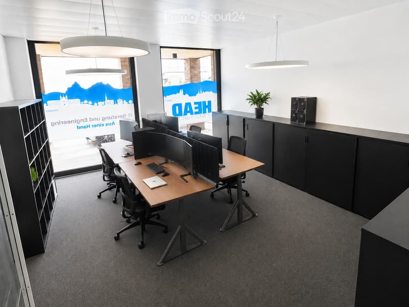Modern office space with a large wooden table, multiple chairs, laptops, keyboards, and computer screens. There are cabinets, a potted plant, and a filing cabinet visible.