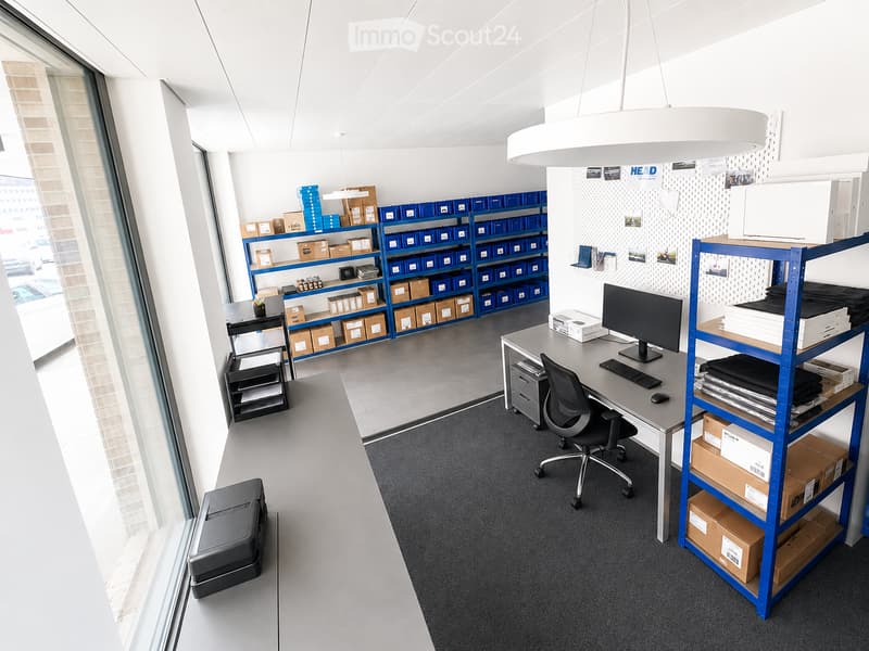 Modern office space with a desk, computer setup, shelves filled with boxes, and a large window providing natural light.