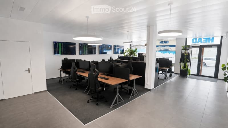 Modern office space with desks, chairs, computer monitors, keyboards, and large screens mounted on the wall. The room features a mix of carpeted and tiled flooring, pendant lights, and a large glass door leading to an outdoor area.
