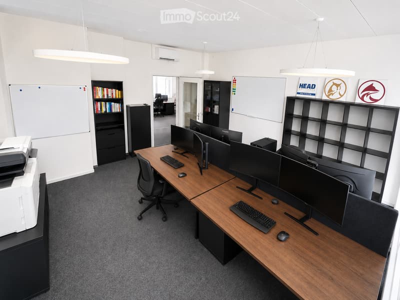 Modern office space with wooden desks, multiple computer monitors, keyboards, mice, ergonomic chairs, a printer, and bookshelves with books and office supplies.