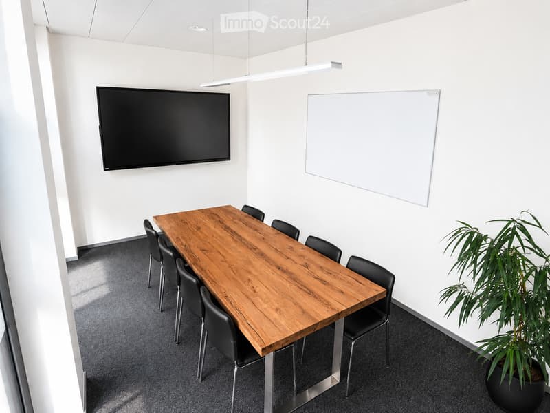 Modern meeting room with a wooden table, black chairs, a large screen, a whiteboard, and a potted plant.