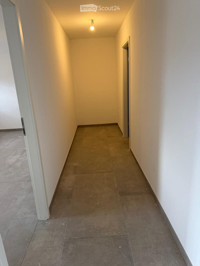 Corridor / Hallway. Empty hallway with tiled floor and white walls, featuring a single light fixture on the ceiling and a partially visible adjacent room on the left.