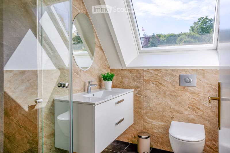 Modern bathroom with beige tiles, white sink, chrome faucet, round mirror, toilet, and small potted plant on the sink counter.