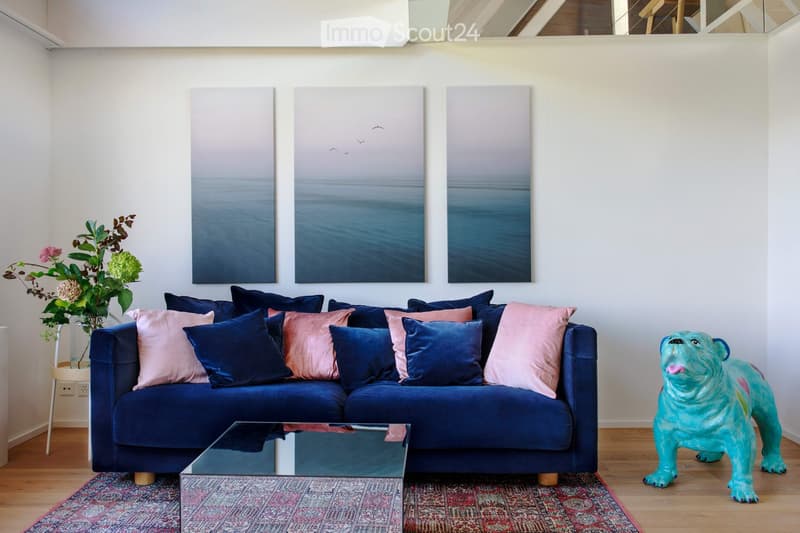 A modern living room with a blue velvet sofa adorned with pink and navy blue throw pillows, a glass coffee table, a colorful rug, a decorative vase with flowers, and a turquoise sculpture of a bulldog.