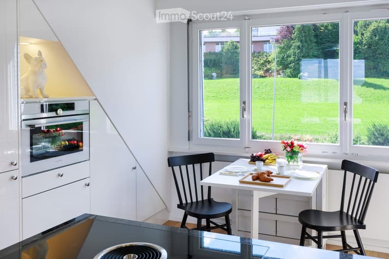 Bright, modern kitchen with white cabinets, a built-in oven, a small dining table with chairs, and a vase of flowers on the table. Large windows offer a view of a green garden.