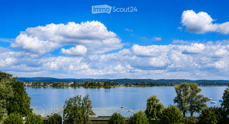 Scenic lakeside view with calm water, distant hills, scattered trees, and a few small boats anchored near the shore.