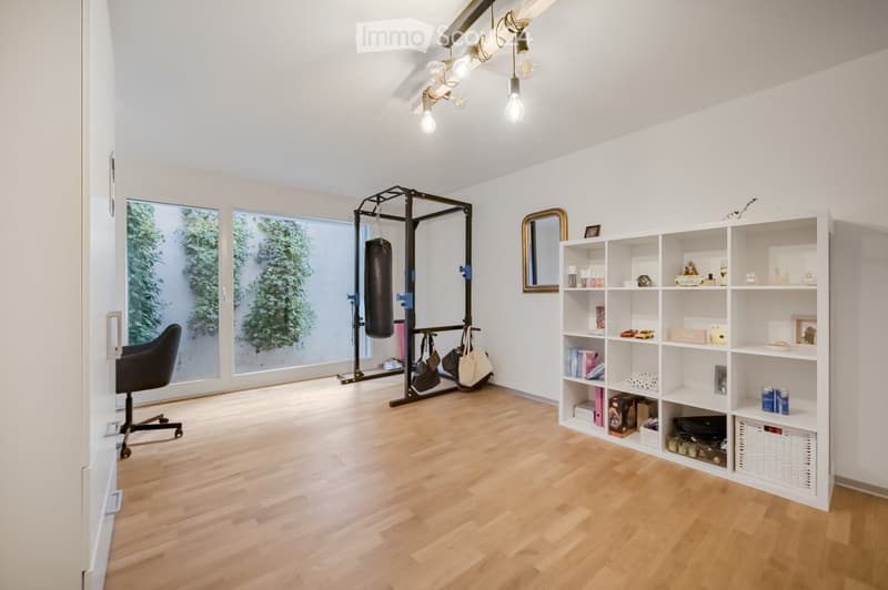 Spacious room with wooden floor, white walls, and a glass door leading to a garden. A punching bag and a gym rack are in the corner. A white shelf holds various items, and a mirror is mounted on the wall. A chair is placed near the door.
