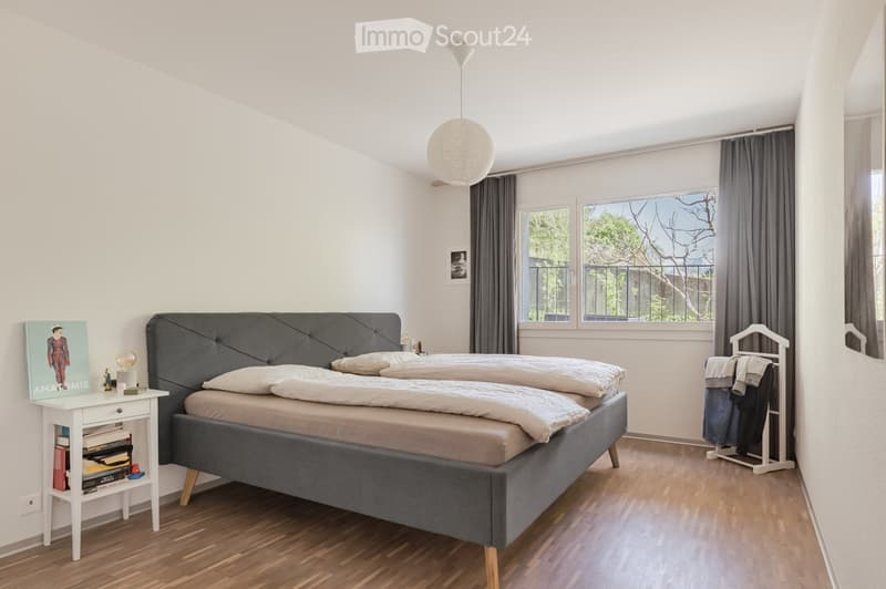 A bedroom with a double bed, nightstand, and a window with curtains. The bed has a gray headboard and wooden legs. The nightstand has a lamp, books, and a poster. The clothes rack is on the right side of the room. The room has a white ceiling and walls, a