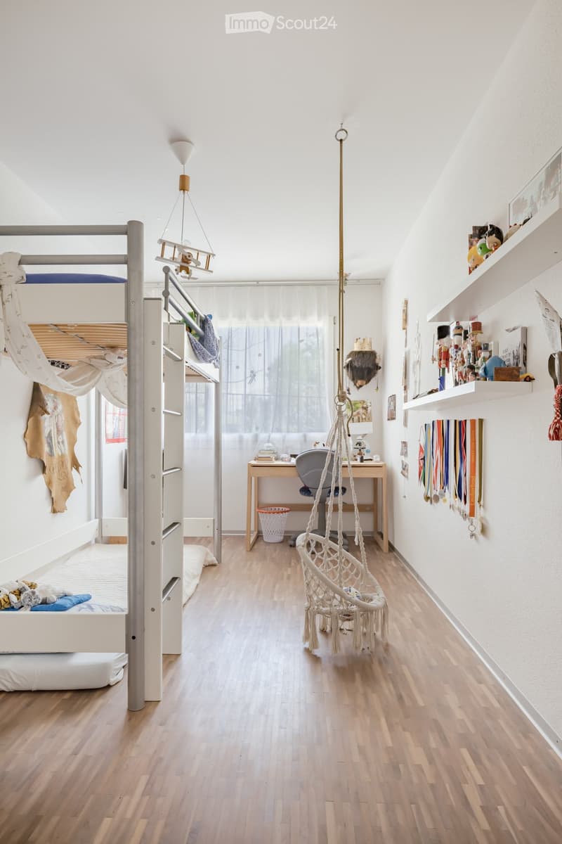 A child's room with a bunk bed, ladder, desk, chair, hammock, basket, shelves, toys, medals, and a lamp.