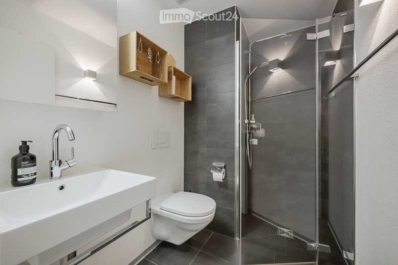 Modern bathroom with white tiles, a white sink, a toilet, and a glass shower enclosure. The sink has a faucet and a soap dispenser. The shower area is equipped with a shower head. The walls are adorned with wall-mounted shelves and light fixtures.