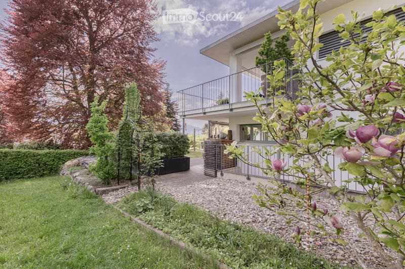 A modern house with a balcony, surrounded by a well-maintained garden with gravel paths, shrubs, and flowering trees.