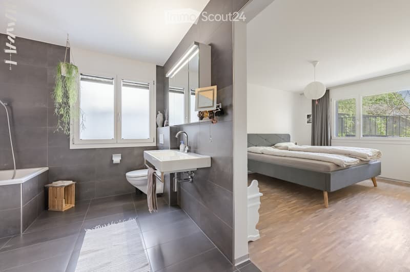 Modern bathroom with gray tiles, white sink, and shower. A wooden stool and a plant add a touch of nature. The bedroom area features a bed with pillows and a lamp.