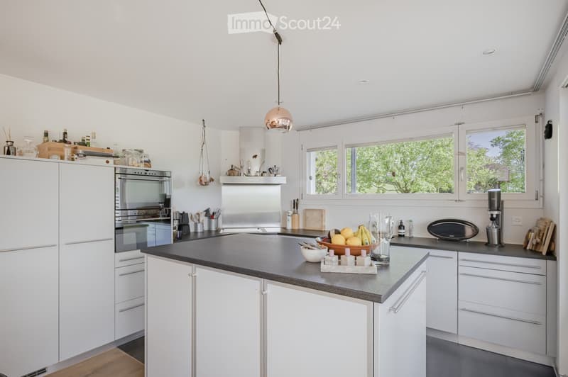 Modern kitchen with white cabinets, gray countertops, and stainless steel appliances. Island with a bowl of fruit and wine glasses. Large windows with a view of trees.