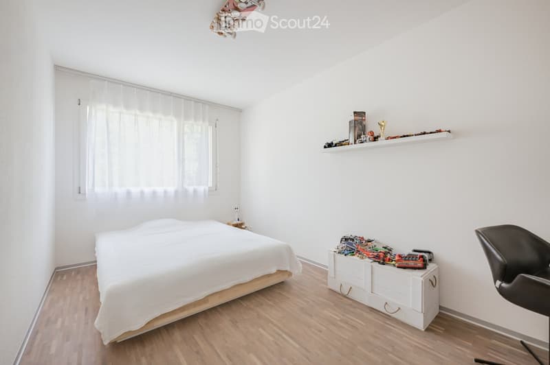 Minimalist bedroom with white walls, wooden floor, bed, nightstand, shelf with toys, and a chair.