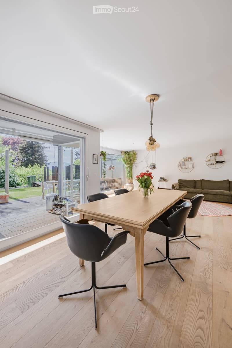 Modern dining area with wooden table, black chairs, and a vase of flowers. The room has large windows leading to a patio.