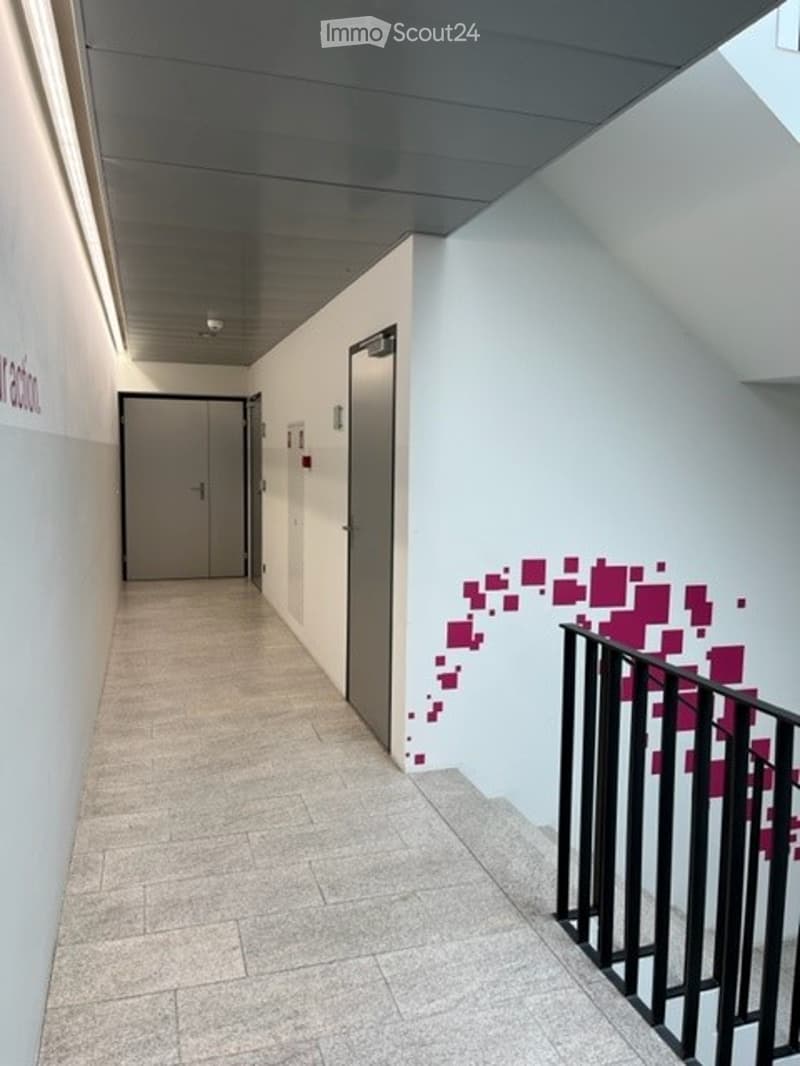 Modern hallway with tiled floor, grey walls, and a staircase leading upwards. Features a black metal railing and a door at the end.