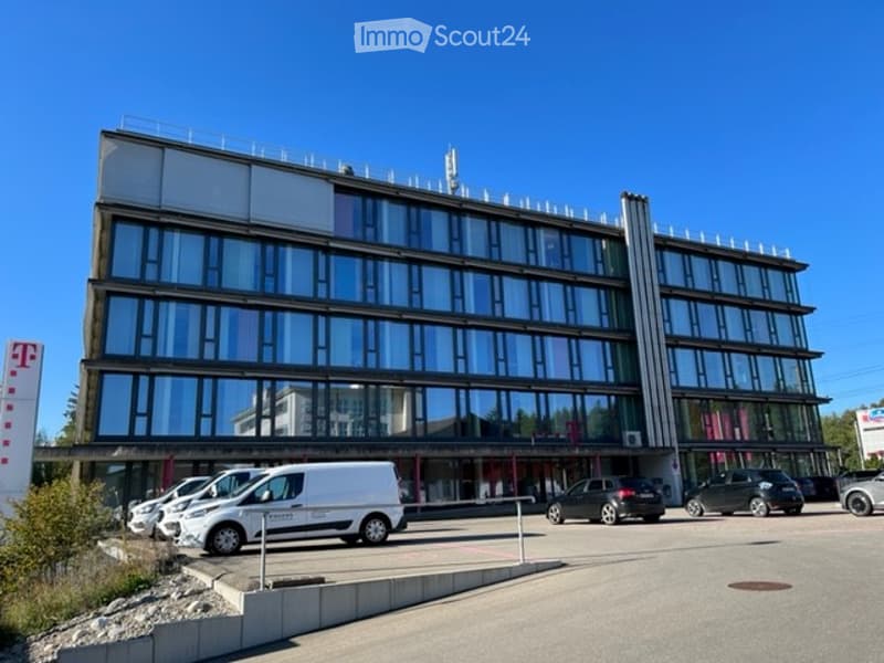 Modern multi-story building with large glass windows and a flat roof, surrounded by a parking area with several cars.