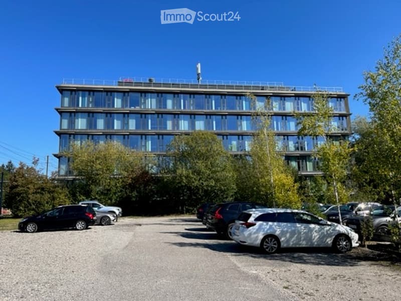 Modern multi-story building with large glass windows and a flat roof, surrounded by trees and a parking lot with several cars.