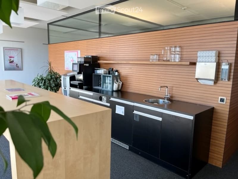 Modern break room with a coffee station, sink, shelves, and a small plant. The walls are wooden panelled, and there is a counter with a coffee machine and cups.