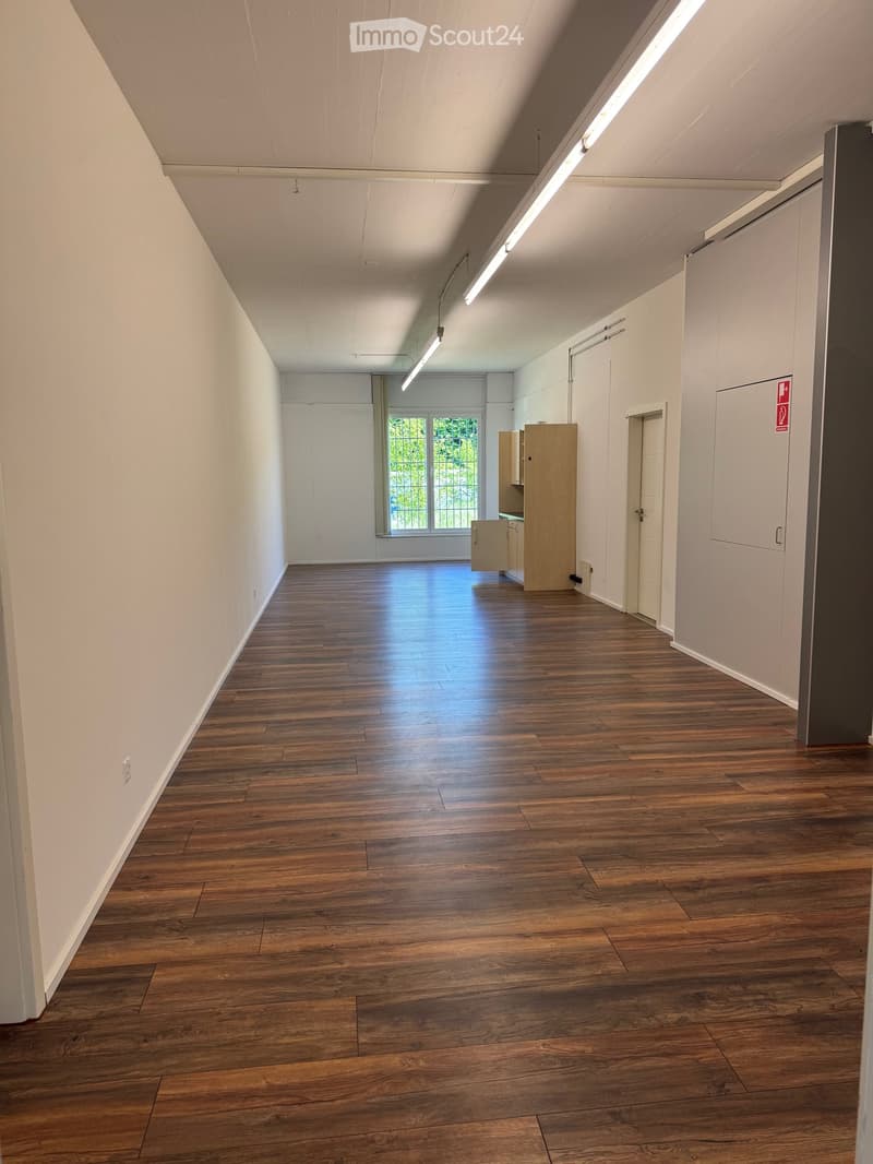 Empty room with wooden flooring, white walls, and a large window at the far end. The space is well-lit with ceiling lights and has a small cabinet and a door visible on the right side.