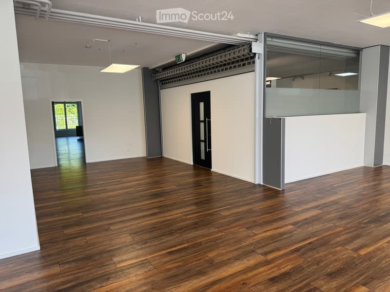 Empty room with wooden flooring, white walls, and large windows allowing natural light. Features include a black door, a glass partition, and exposed ceiling beams.