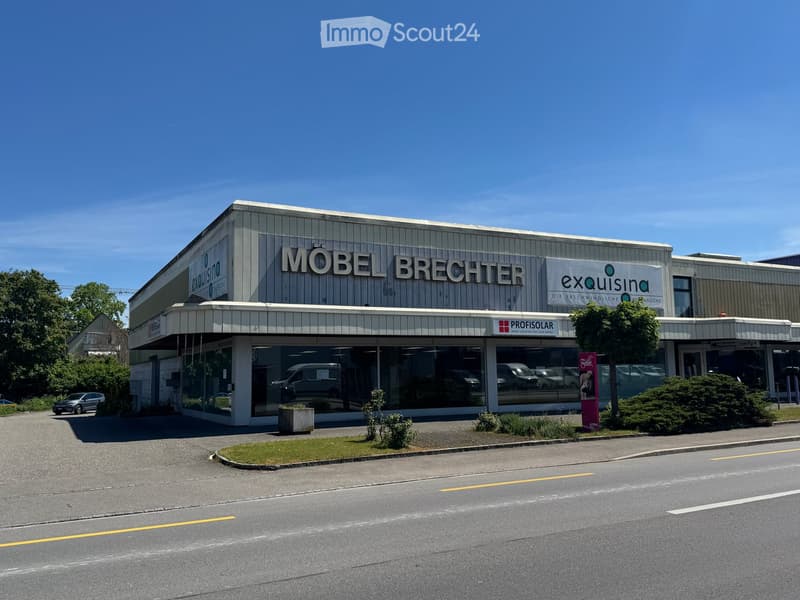 Commercial building with signage reading "MÖBEL BRECHTER" and "exquisina. DIE ERSCHWINGLICHE KUCHENKOCHE" on the facade, large glass windows, and a parking area in front.