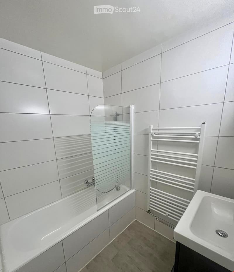 Modern bathroom with white tiled walls and floor, featuring a bathtub with a glass shower enclosure, a white radiator, and a sink.