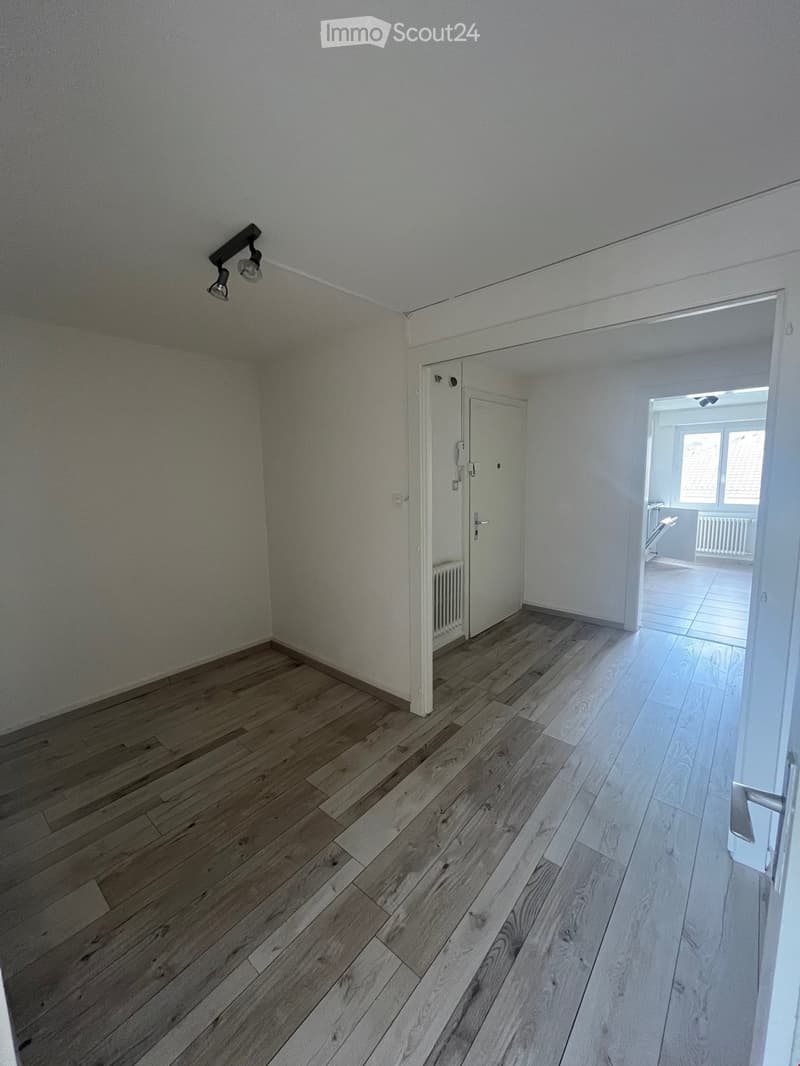 Empty room with wooden flooring, white walls, and a door leading to another room. The space is unfurnished and appears to be a hallway or entryway.