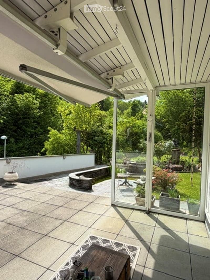 Balcony / Terrace. Modern covered outdoor terrace with large glass sliding doors, tiled floor, and a view of a lush green garden.