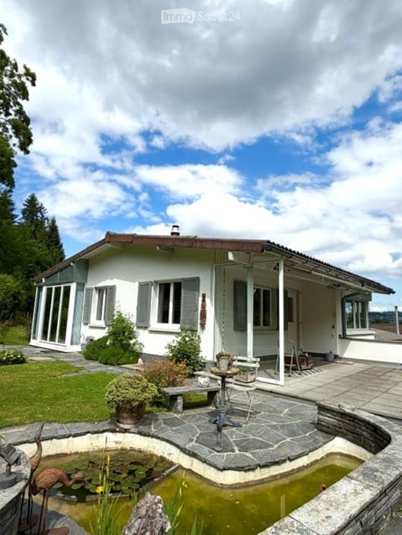 House. Single-story white house with grey shutters, surrounded by a well-maintained garden, featuring a small pond with lily pads, a stone bench, and decorative items.