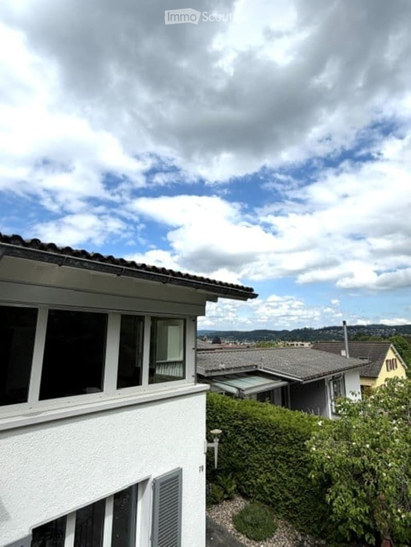 Building. View of a residential building with white walls, large windows, and a sloped roof, surrounded by greenery and neighboring houses.