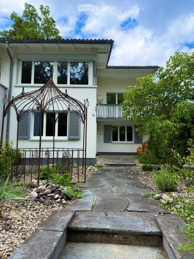 House. Two-story white house with multiple windows, shutters, and a balcony. A decorative metal gazebo is positioned in the front garden, and a stone pathway leads to the entrance.