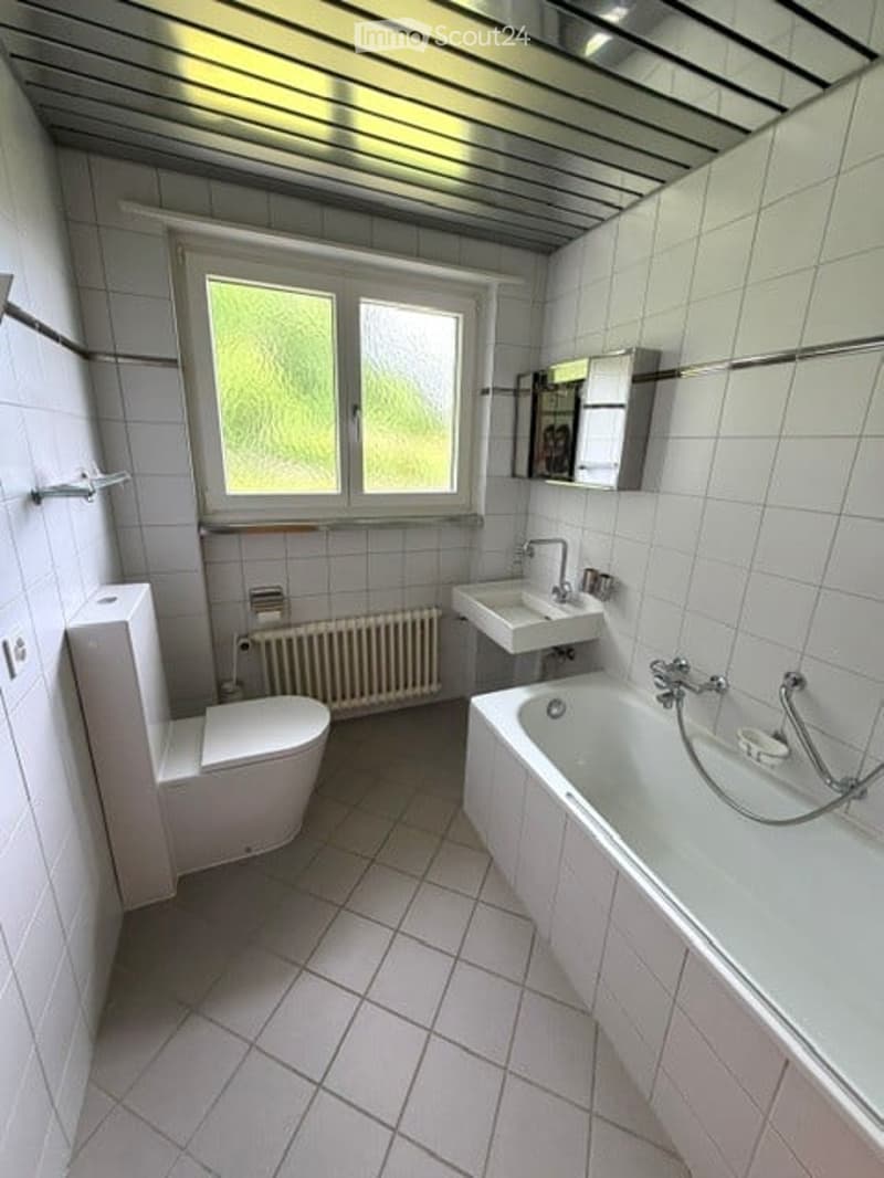 Bathroom. Bright bathroom with white tiles, a toilet, a bathtub, a sink, a radiator, and a mirror. The window offers a view of greenery outside.