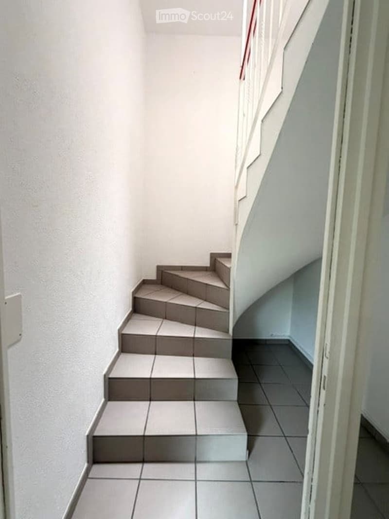 Corridor / Hallway. A narrow, tiled hallway with a staircase leading upwards, featuring white walls and a simple wooden handrail.