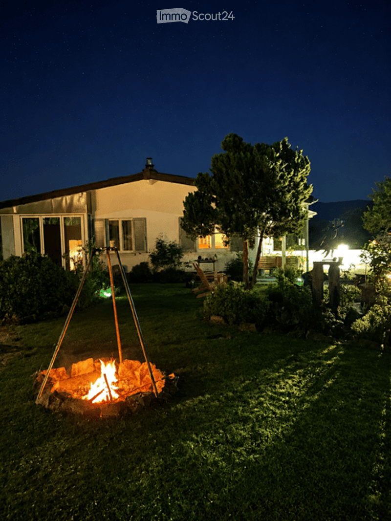 Garden / Outdoor Area. Nighttime view of a house with a lit fire pit in the front yard, surrounded by trees and shrubs.