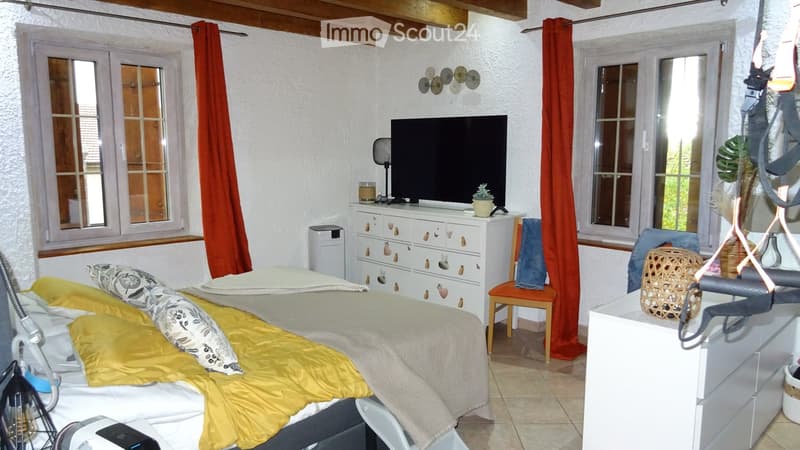 Bedroom with bed, white walls, wooden ceiling, two windows, TV on top of a white cabinet, wooden chair, red curtains, and decorative items.