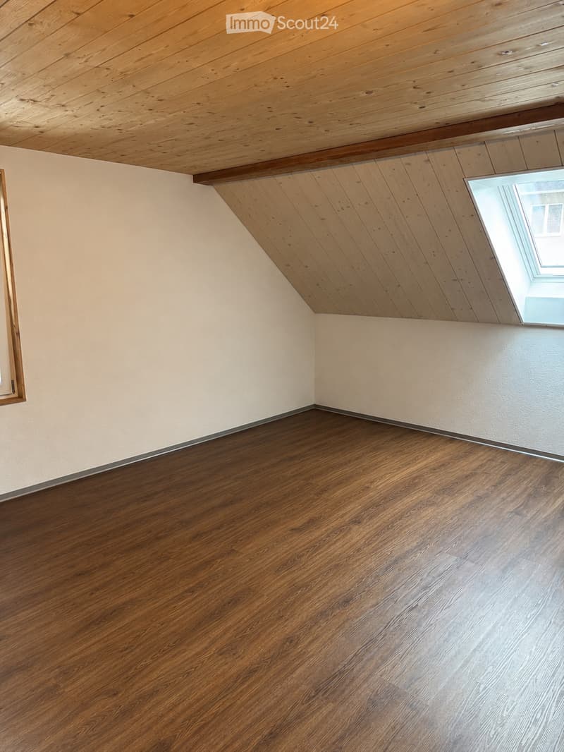 Empty room with wooden flooring and light-colored walls, featuring a skylight window and exposed wooden ceiling beams.