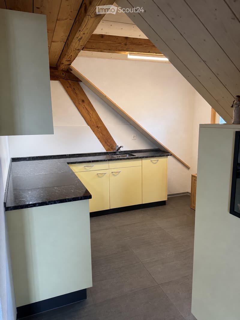 Modern kitchen with light-colored cabinets, a dark countertop, and a sink under a window. The room features exposed wooden beams on the ceiling and a wooden floor.