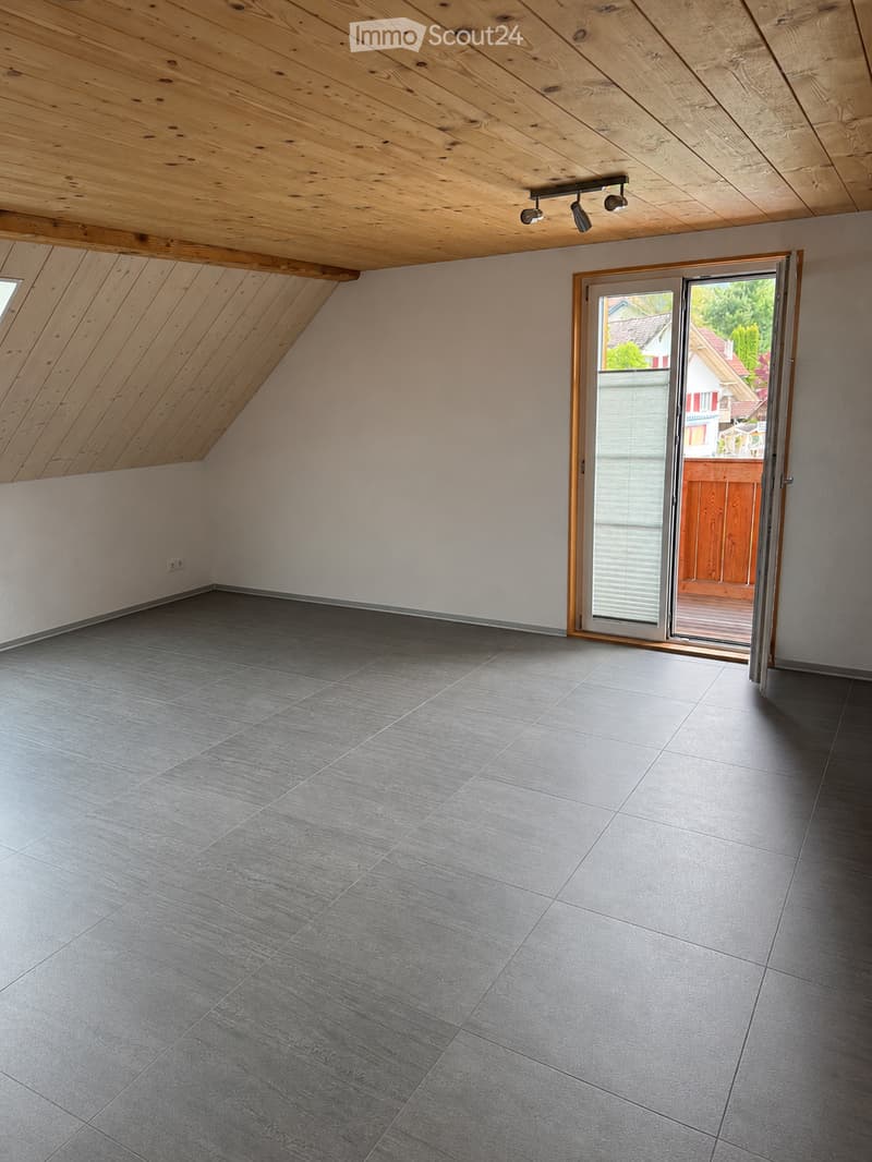 An empty attic room with wooden slanted ceilings, grey tiled flooring, and a large sliding glass door leading to an outdoor deck.
