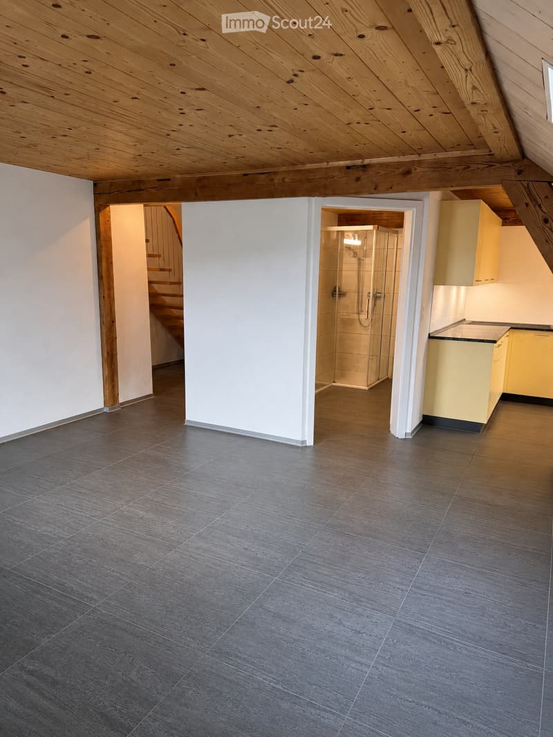 An empty room with a wooden ceiling, grey tiled floor, and visible doorways leading to a staircase and a bathroom area with a glass shower enclosure.