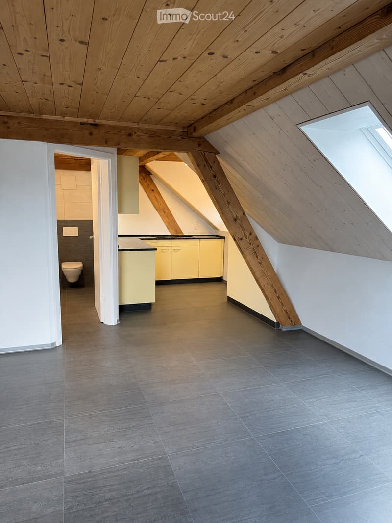 Modern attic space with wooden beams, grey tiled floor, cream-colored cabinets, and a small bathroom area with a toilet visible.