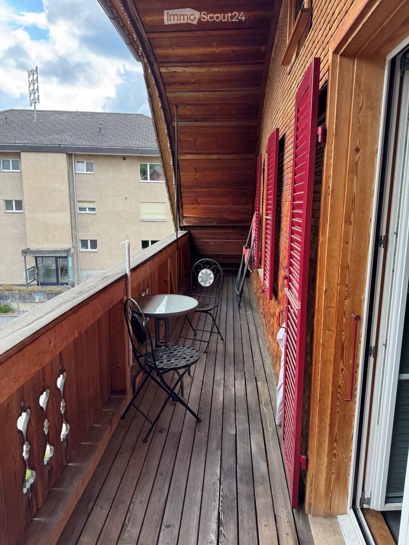 Wooden balcony with a small round table and two black metal chairs, red shutters, and a view of a neighboring building.