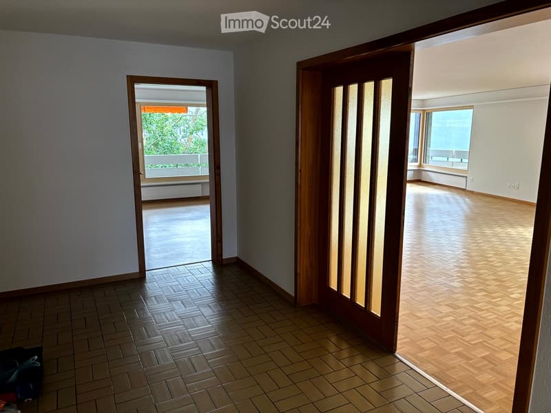 Empty room with tiled floor, wooden doors, and access to a balcony area with wooden flooring and large windows.