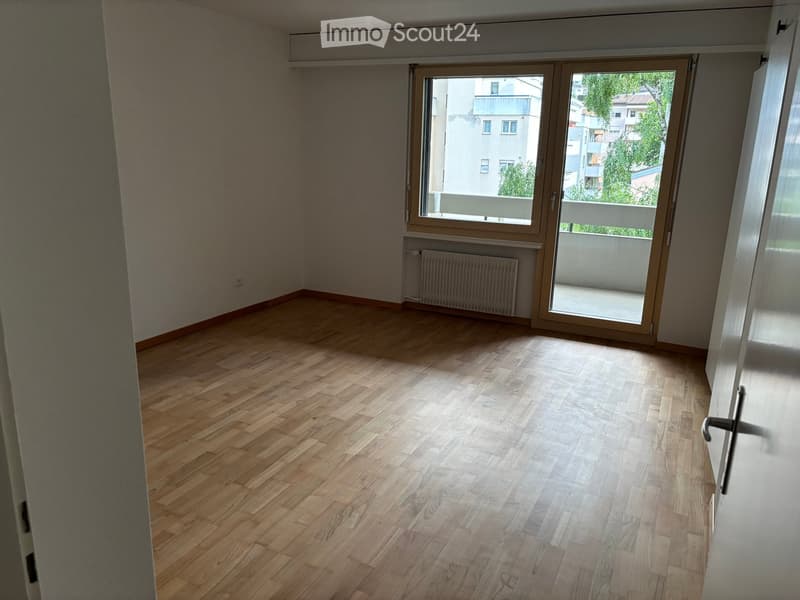 Empty room with wooden flooring, large window with sliding doors leading to a balcony, radiator, and visible electrical outlet.