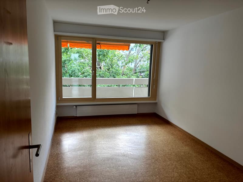Empty room with tiled floor, white walls, large window with view of greenery and balcony, radiator below window.