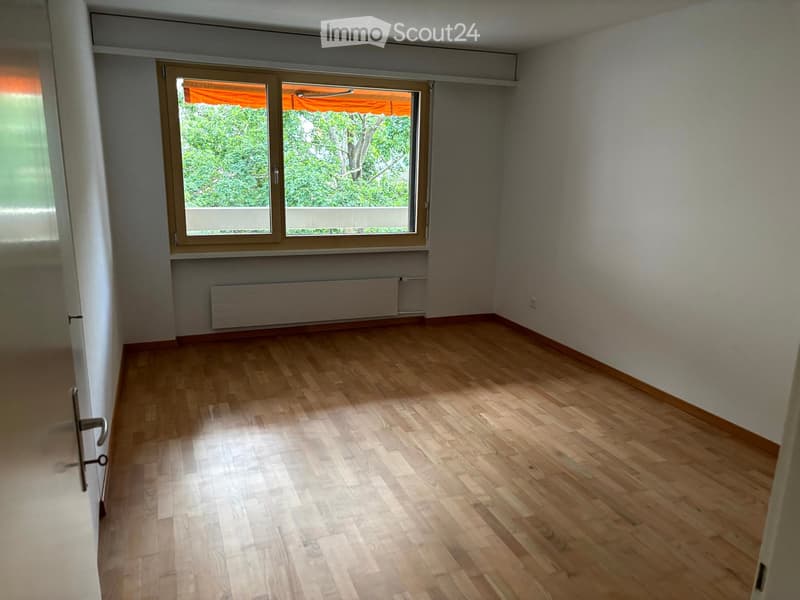 Empty room with wooden flooring, white walls, large window with blinds, radiator, and a door visible on the left side.