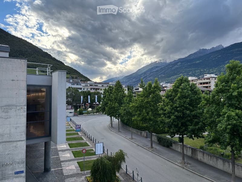 Modern building entrance with glass facade, surrounded by landscaped area with trees, grass patches, and distant mountain view.