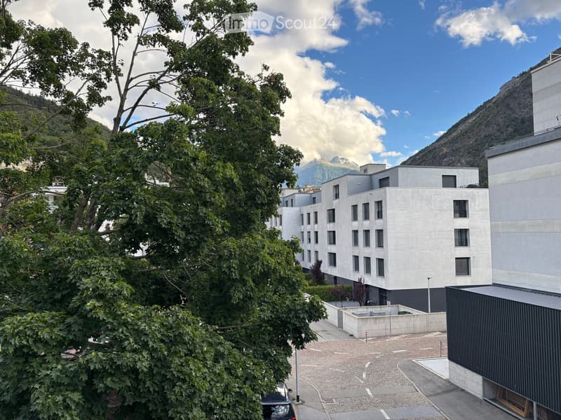 Modern multi-story building with clean lines and large windows, surrounded by trees and a paved courtyard, with a mountain view in the background.